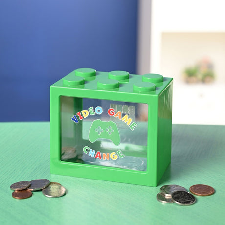 Green plastic building block money box with six studs and a clear window reading "Video Game Change" around a game controller icon, shown with scattered coins on a green surface.