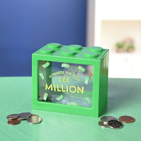 Green plastic building block money box with six top studs and a clear front window reading "Towards the first £££ million" amid coins on a green surface.
