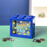 Navy blue plastic money box shaped like a building block with six top studs and a clear front window reading "Bank of Dad" amid printed gold coins, shown with loose coins on a green surface.
