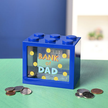 Navy blue plastic money box shaped like a building block with six top studs and a clear front window reading "Bank of Dad" amid printed gold coins, shown with loose coins on a green surface.