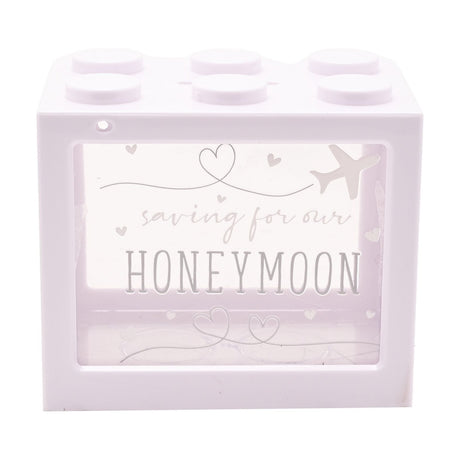White plastic brick-shaped money box with six round studs and a clear window reading "saving for our HONEYMOON" with heart and aeroplane line art, on a white background.