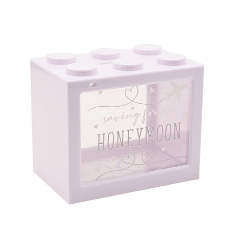 White plastic money box shaped like a building brick with six top studs and a clear front window printed "saving for our HONEYMOON" with hearts and an aeroplane design.