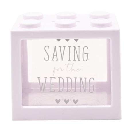 White plastic building block money box with six top studs and clear front panel reading "SAVING for the WEDDING" in grey text with small hearts, on a plain white background.