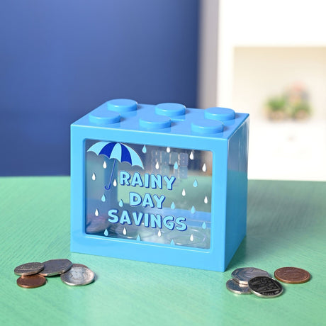 Blue plastic building block money box with six top studs and a clear front window reading "Rainy Day Savings" beside an umbrella and raindrop design, shown with scattered coins on a green surface.