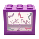 Purple plastic brick-shaped money box with six top studs and a clear front window printed "SHOE FUND" amid lilac, pink and grey stiletto heel graphics, shown on a white background.