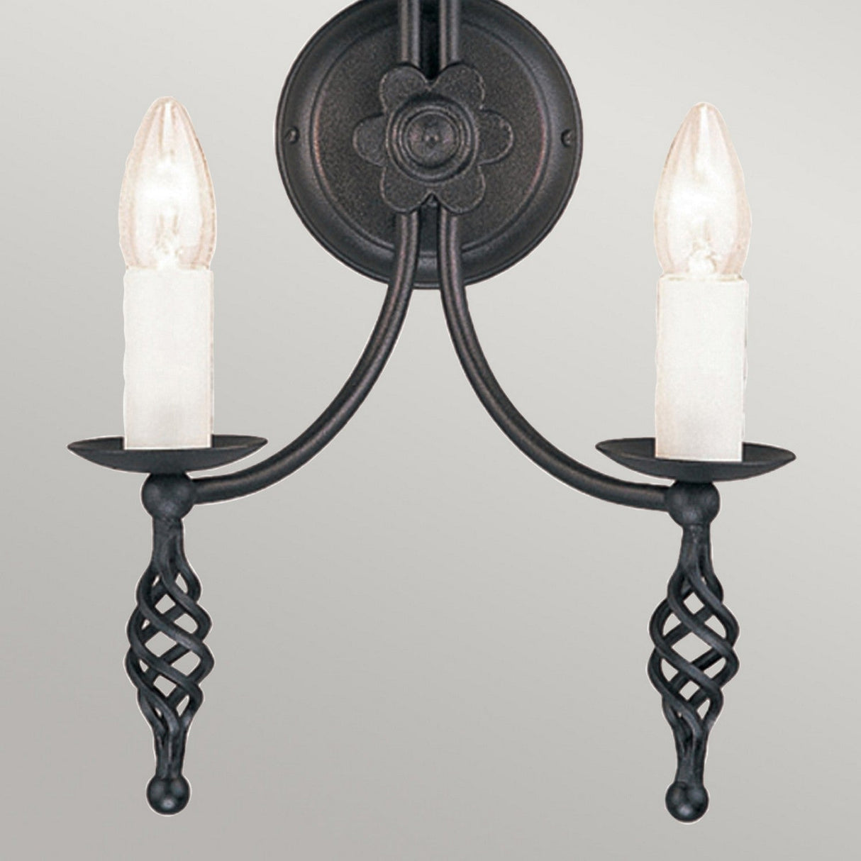 Introducing the Belfry 2 Light Wall Light - Black, a stunning piece of medieval artistry featuring candle-shaped bulbs and intricately twisted decorative accents. This hand-forged wrought iron sconce is beautifully mounted on a circular backplate adorned with a floral design, offering an elegant contrast against any neutral backdrop.