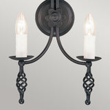 Introducing the Belfry 2 Light Wall Light - Black, a stunning piece of medieval artistry featuring candle-shaped bulbs and intricately twisted decorative accents. This hand-forged wrought iron sconce is beautifully mounted on a circular backplate adorned with a floral design, offering an elegant contrast against any neutral backdrop.