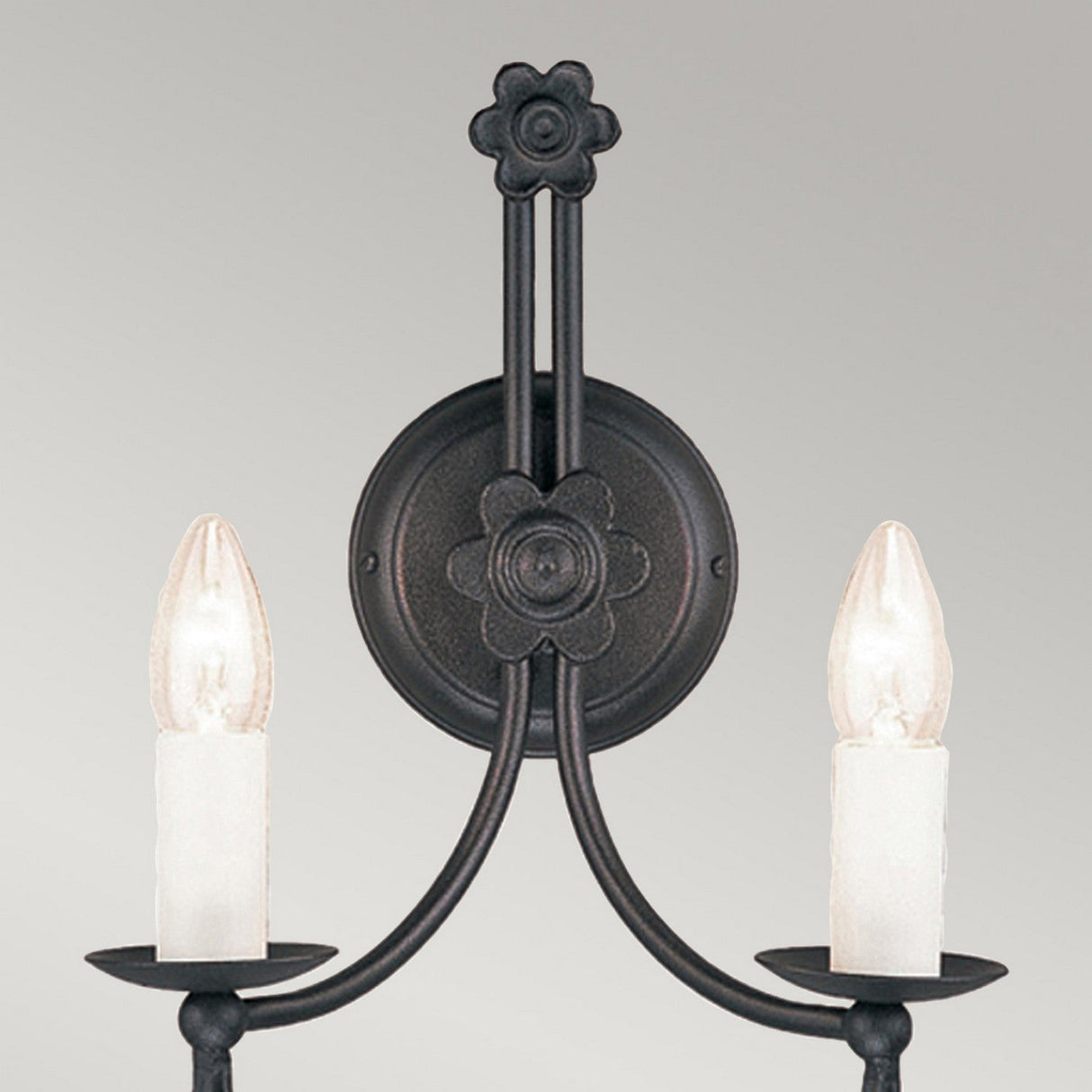 The Belfry 2 Light Wall Light in black displays medieval artistry with its round base and floral accents. Featuring two candle-shaped light bulbs on gracefully curved arms, this hand-forged metal sconce stands out against a light gray wall, bringing timeless elegance to any room.