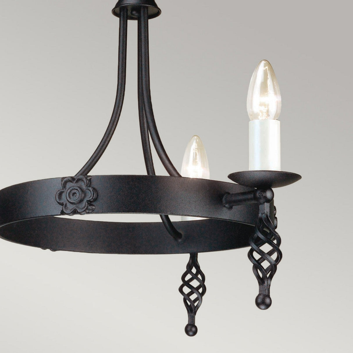 The Belfry 3 Light Chandelier - Black highlights stunning medieval artistry with a hand-forged metal frame. This elegant black chandelier displays two candle-shaped light bulbs and intricate twisted metal designs featuring a floral motif, beautifully contrasted against a simple light gray background.