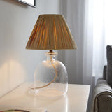 Caldwood 25cm tapered lamp shade in natural raffia atop a textured glass lamp base, shown lit on a bedside table in a living room setting.