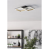 Calliera LED flush ceiling light with two overlapping square frames in black and brass, mounted on a light grey ceiling above a living room shelving unit and plant.