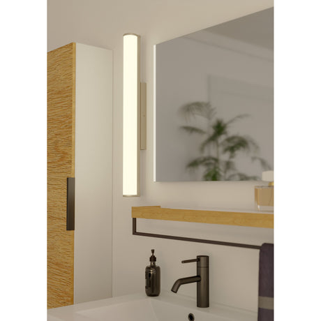 Calnova LED bathroom wall light in satin nickel, a slim frosted glass bar fixture lit beside a bathroom mirror above a basin with dark tap and soap dispenser.