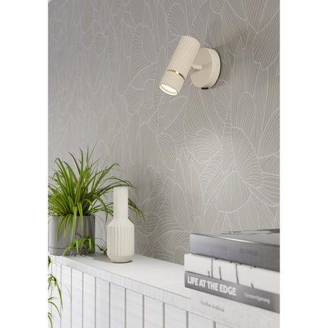 Caminia Wall Light in sand with a brushed brass ring, fluted cylindrical head on a round steel plate, shown lit on patterned wallpaper above a shelf with books and plants.