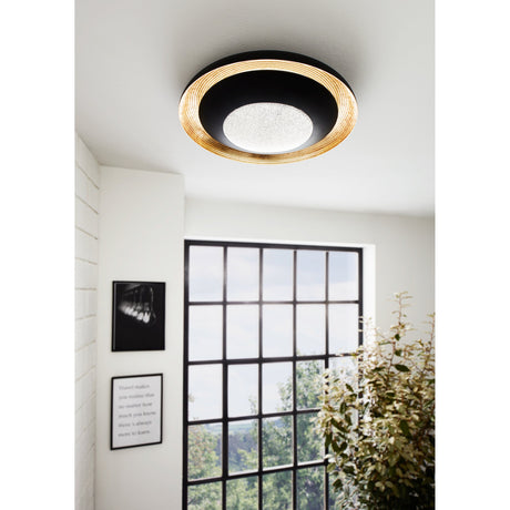 Canicosa 2 LED flush ceiling light in black and gold, round ribbed rim with sparkling centre diffuser, shown installed on a hallway ceiling near a window and plant.