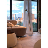 A cozy living room corner features a curved beige sofa with a soft throw, pillow, and open book. Ambient light comes from the Capuccina LED Floor Lamp 8.5W 3000K - Black, while a large window reveals a cityscape at sunset.