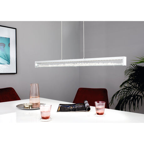 Cardito 1 LED pendant light with a slim chrome bar and clear glass channel filled with sparkling glass pieces, suspended above a white dining table in a grey room.