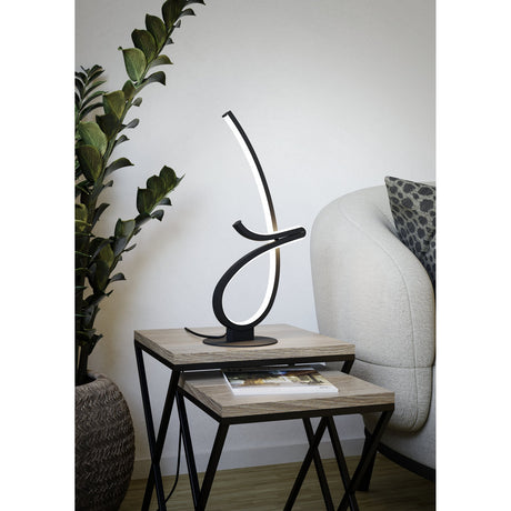 Cardoncito LED Table Lamp in black steel, its curved illuminated ribbon shape set on a round base atop a wooden side table beside a sofa and potted plant.