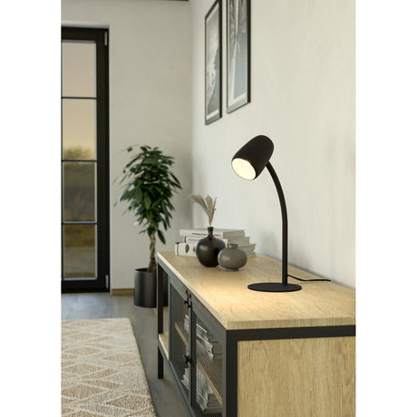 Carlo Table Lamp in matt black steel with a curved gooseneck stem and domed shade, shown lit on a wooden sideboard in a living room setting.