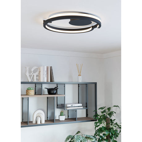 Carmanne LED Flush Ceiling Light in black, an overlapping ring design with white diffuser band, shown mounted on a living room ceiling above a shelving unit with books and decor.