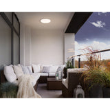 Castagneto 31cm round LED flush ceiling light in white, shown illuminating a covered balcony seating area with rattan sofas and cushions.