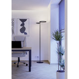 Castelpoto 1 LED floor lamp in black with slim pole and two offset illuminated rings, shown lit beside a desk and chair in a home office setting.