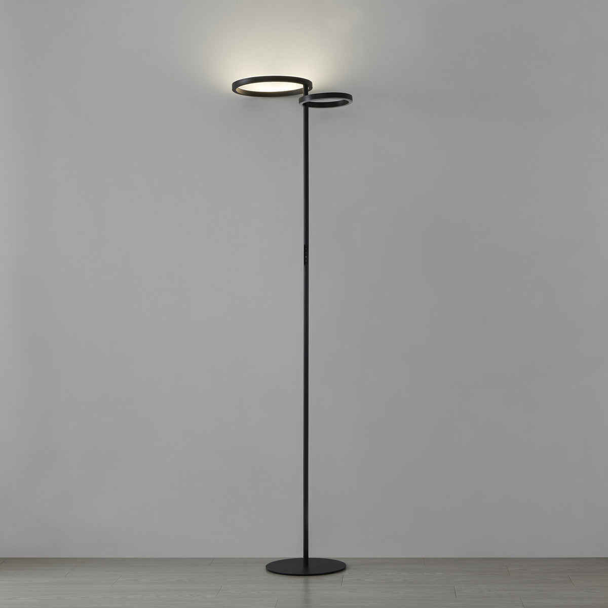 Castelpoto 1 LED floor lamp in black, tall slim pole on a round base with two offset ring-shaped shades glowing warm white, set against a plain grey background.