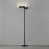 Castelpoto 1 LED floor lamp in black, tall slim pole on a round base with two offset ring-shaped shades glowing warm white, set against a plain grey background.