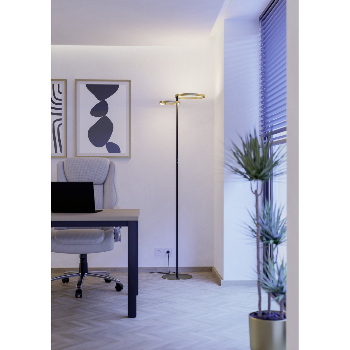 Castelpoto 1 LED floor lamp with two brushed brass rings atop a slim black steel stem and round brushed brass base, shown beside a desk in a home office setting.