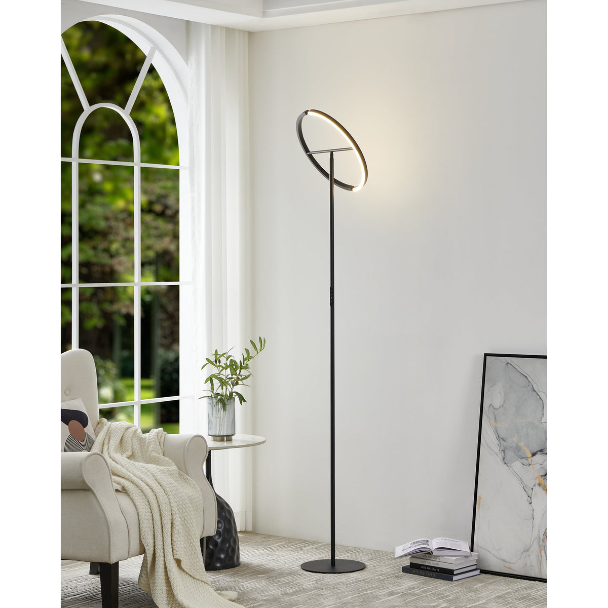 Castelpoto LED floor lamp in black steel with a tilted ring-shaped light head on a slim stem and round base, shown beside a reading chair and window in a living room.