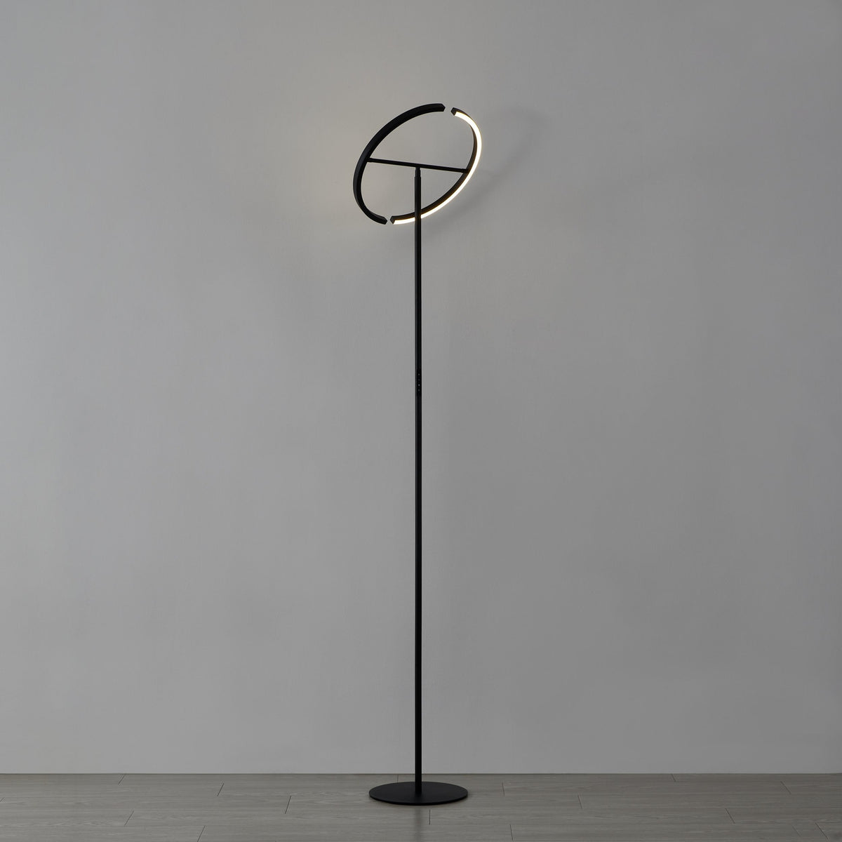 Black steel Castelpoto LED floor lamp with tall slim stem, round base, and angled split-ring top emitting warm white light, shown on a plain grey background.