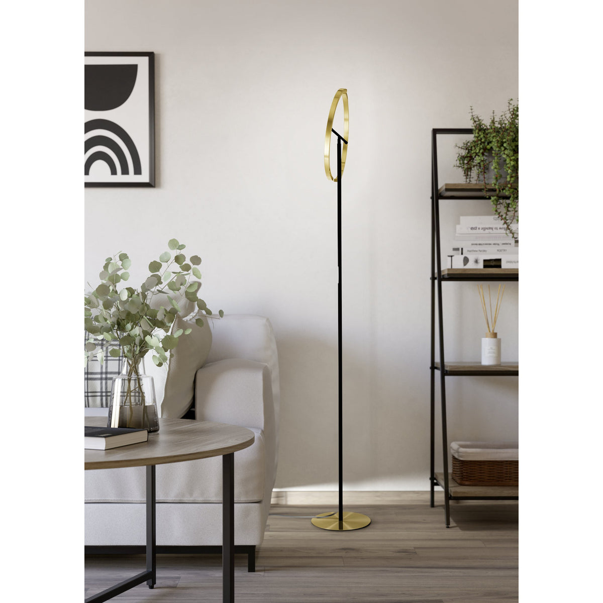 Castelpoto LED floor lamp with slim black steel stem and split ring head in brushed brass, round brass base, shown in a living room beside a sofa and shelving.