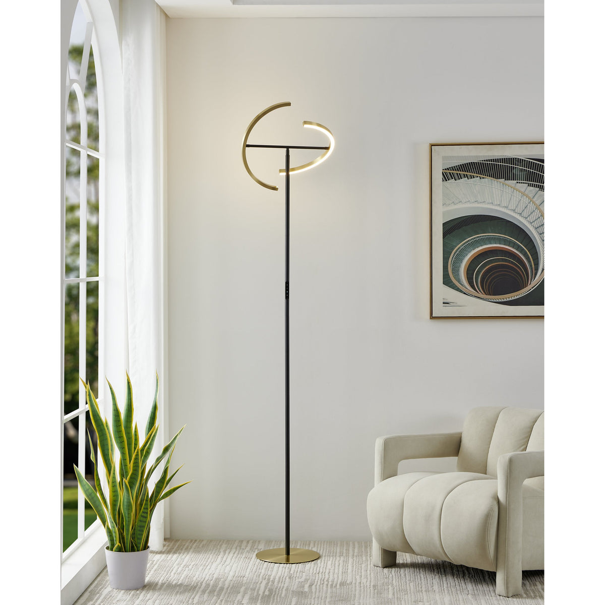 Castelpoto LED Floor Lamp with a slim black steel stem, split-ring brushed brass head and round brushed brass base, shown lit in a living room beside a window and armchair.