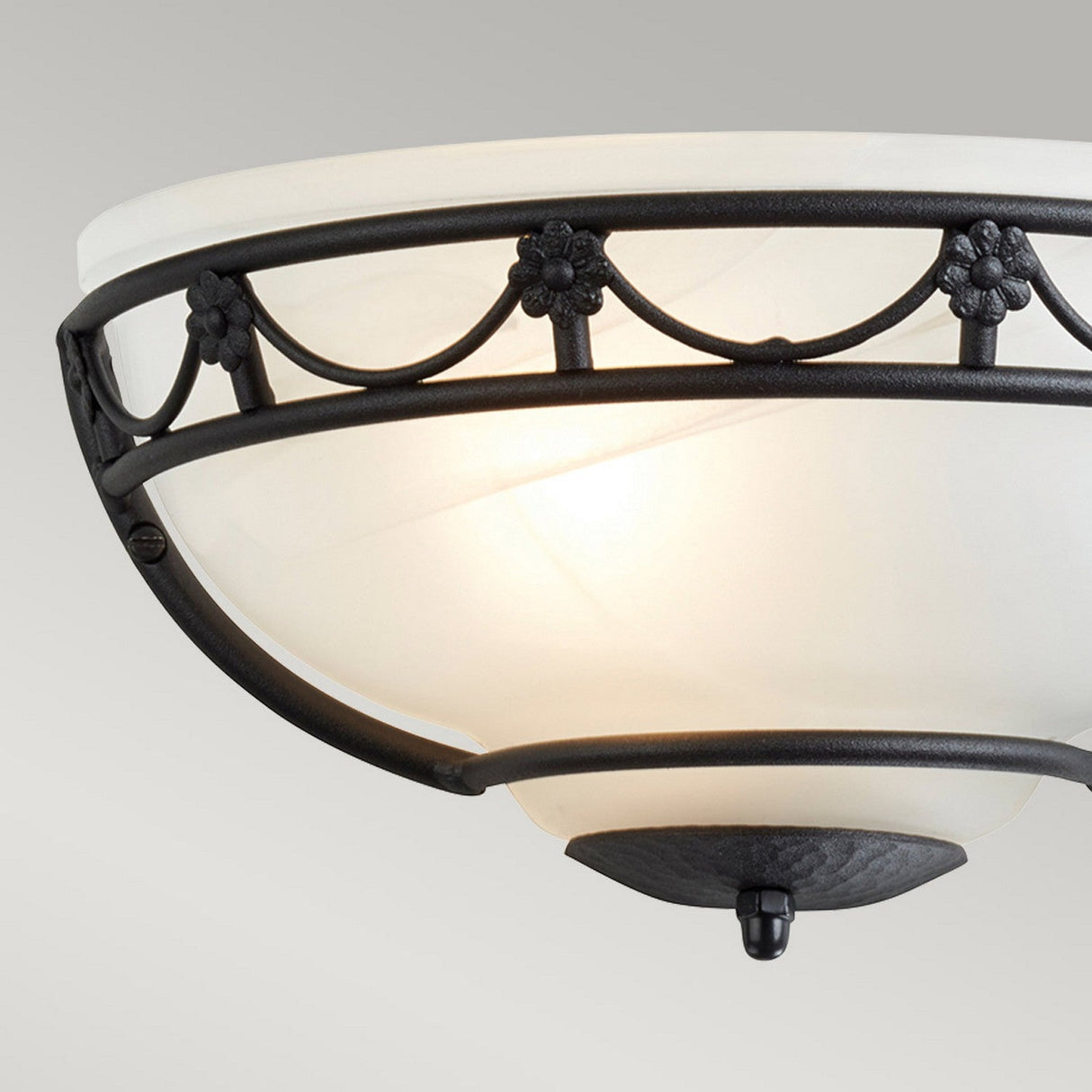 A close-up of the Carisbrooke Wall Uplighter - Black showcases its alabaster-style glass shade encased in a black metal frame. The Gothic-inspired design highlights floral accents and scalloped detailing, all set against a neutral gray background.