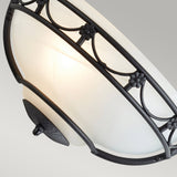 A close-up showcases the Carisbrooke Wall Uplighter - Black, complete with an alabaster-style glass shade. Its frame displays intricate black metalwork adorned with floral details, while a warm light from within accentuates the vintage sophistication of its design against a neutral backdrop.