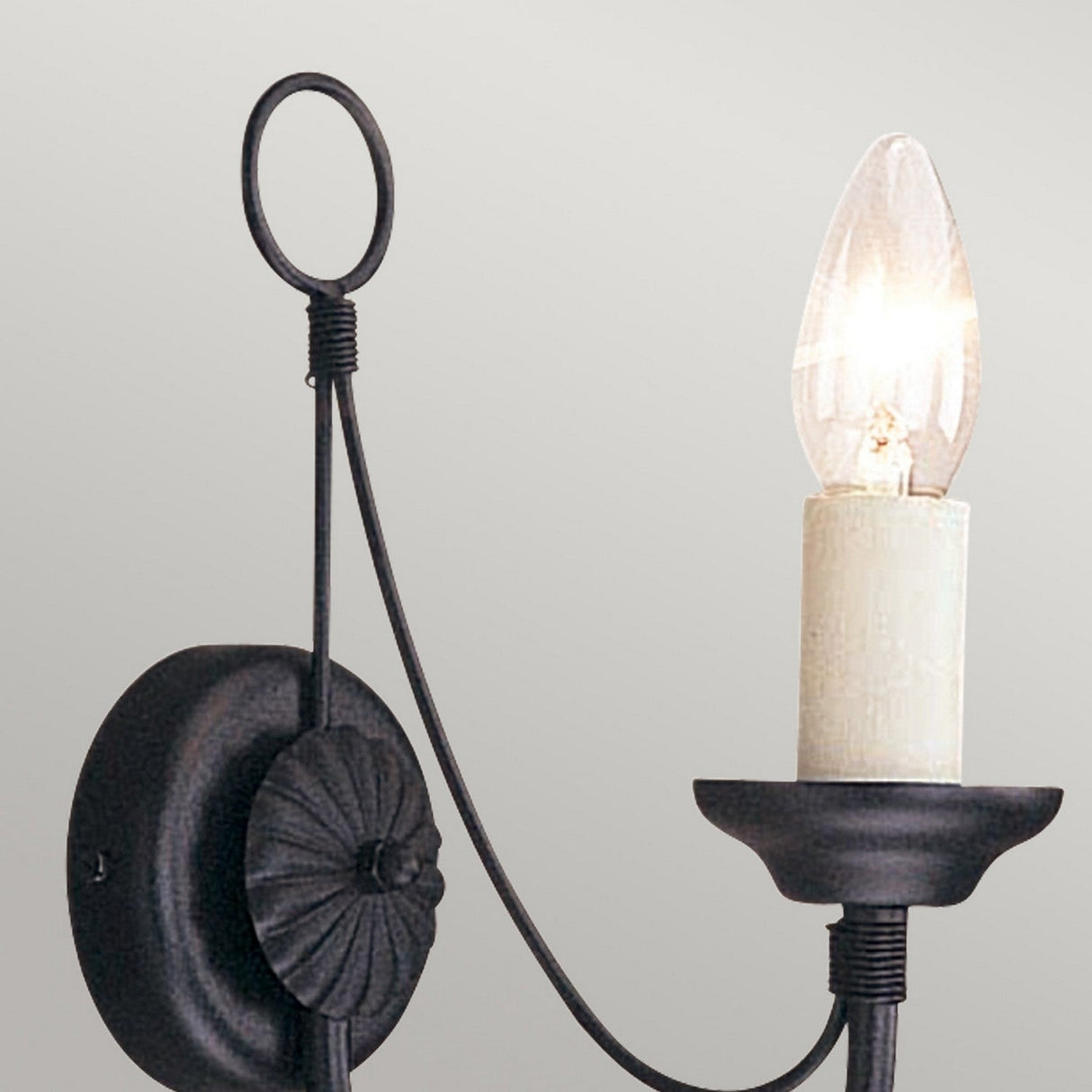 The Carisbrooke Wall Light - Black is a wrought iron wall sconce with a circular base and decorative loop at the top, offering a vintage feel with its exposed bulb that resembles a candle flame. When illuminated, the bulb casts a warm glow against the plain light gray wall.