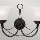 The Carisbrooke 2 Light Wall Light - Black is a wrought iron fixture featuring two upward-facing frosted glass lamps that exude vintage charm. Its design showcases gracefully curved metal arms and a central decorative rosette at the base, elegantly contrasted against a simple, muted gray background.