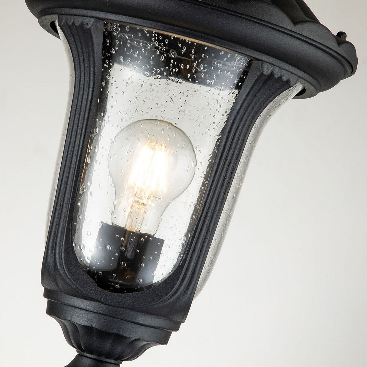 Close-up of the Chicago Small Pillar Lantern - Black, showcasing its vintage-style design with a curved black frame and clear glass casing. Inside, a glowing filament bulb embodies classic outdoor lighting charm as droplets of water shimmer on the glass surface.
