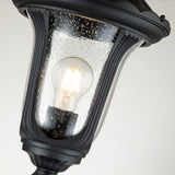 Close-up of the Chicago Small Pillar Lantern - Black, showcasing its vintage-style design with a curved black frame and clear glass casing. Inside, a glowing filament bulb embodies classic outdoor lighting charm as droplets of water shimmer on the glass surface.