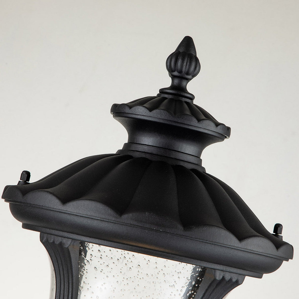 Close-up of a decorative black lantern top for outdoor use. The Chicago Small Pillar Lantern - Black showcases an ornate finial and a scalloped edge. Its textured glass, speckled with droplets, enhances the vintage aesthetics against a neutral background, adding classic charm to any outdoor lighting arrangement.