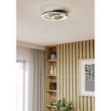 Cembrano LED flush ceiling light in black and natural wood with two overlapping illuminated rings, mounted above a wood-panelled wall with shelving in a living space.