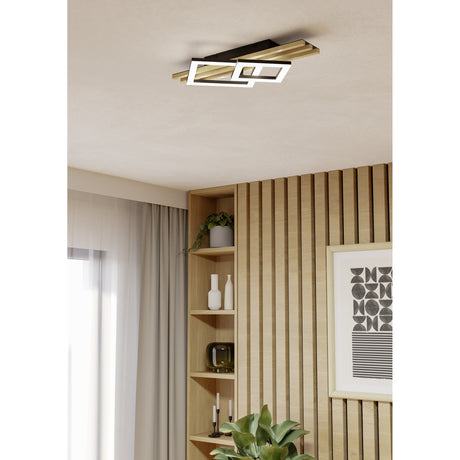 Cembrano LED flush ceiling light in black and natural wood, mounted on a living room ceiling above a wood-slatted wall with shelving and decor.