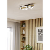 Cembrano 50cm LED flush ceiling light in black and natural wood, with two overlapping rings of varying size and white diffuser faces, mounted in a wood-panelled living room.
