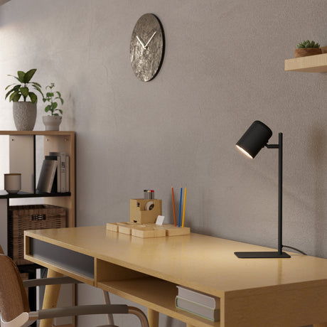 Ceppino Table Lamp in matt black steel with a white-lined cylindrical head on a slim stem, shown lit on a wooden desk beside a clock and shelves.
