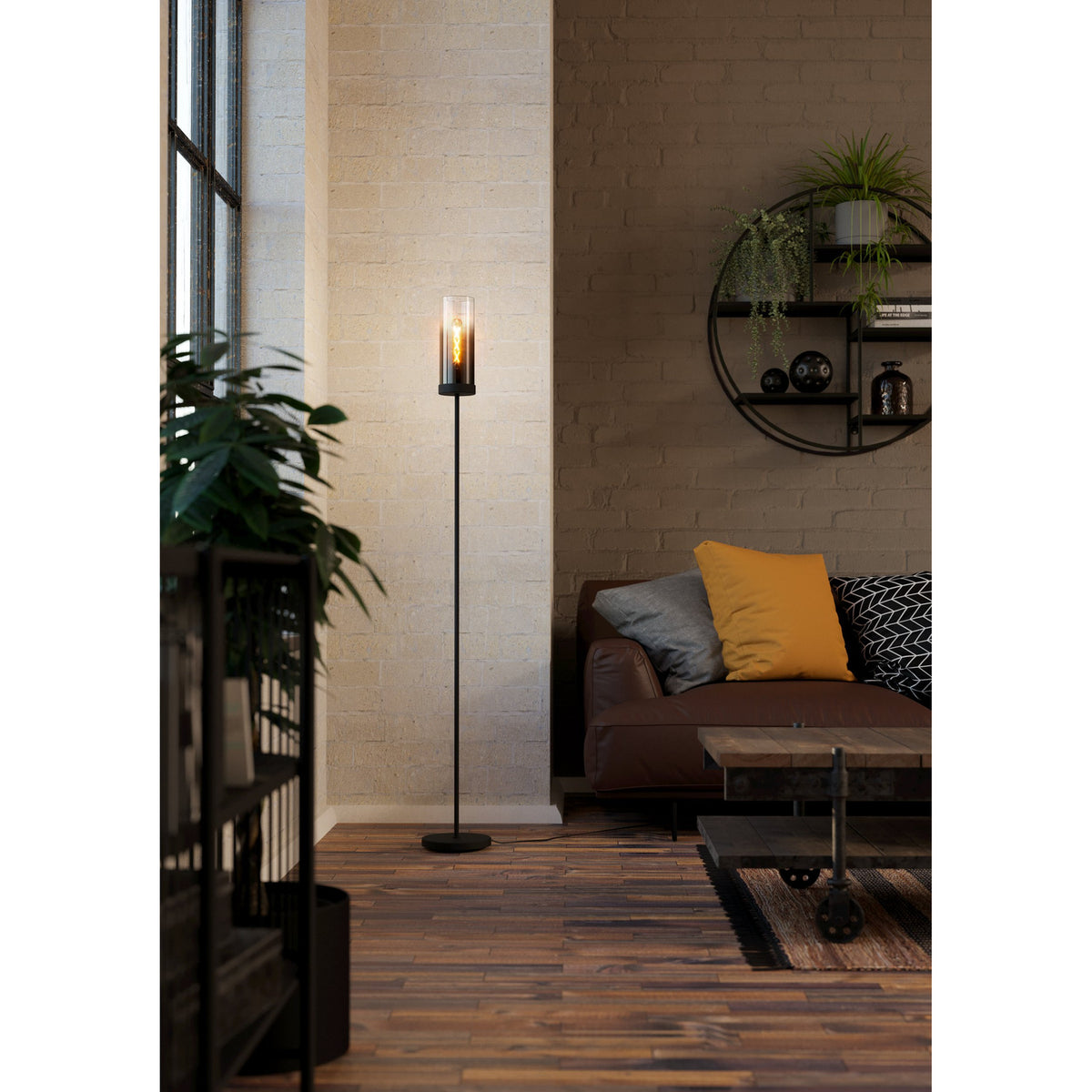 Cesarne Floor Lamp in black steel with a slim column and smoked glass cylinder shade, shown lit beside a sofa in a living room setting.