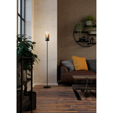 Cesarne Floor Lamp in black steel with a slim column and smoked glass cylinder shade, shown lit beside a sofa in a living room setting.
