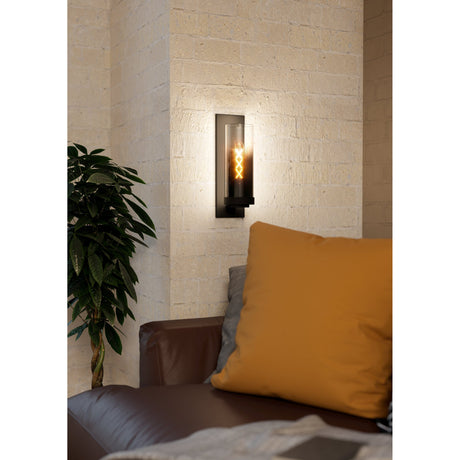 Cesarne Wall Light in black, a tall steel wall fixture with smoked glass cylinder, shown illuminated on a stone wall beside a leather sofa and mustard cushion.