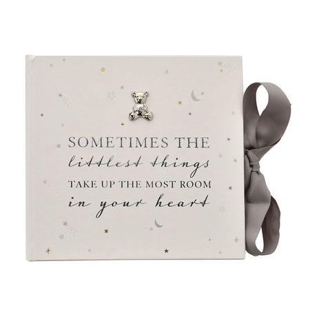 White square baby photo album for 50 6x4in prints, with foiled "Sometimes the littlest things..." script, a silver teddy bear charm, star and moon accents, tied with a grey ribbon bow.