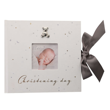 White christening day photo album with star and moon print, silver-tone teddy bear, square photo window showing a sleeping baby, gold "Christening day" script, and grey ribbon tie.