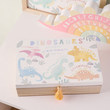 Rectangular wooden keepsake box with white lid illustrated with five named dinosaurs in pastel colours, tan wood base, and a yellow tassel on the front catch.