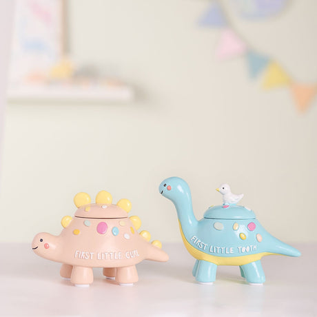 Resin dinosaur keepsake box set with lift-off lids, blue "First Little Tooth" dinosaur with bird finial and pink "First Little Curl" stegosaurus with yellow plates, shown on a tabletop.